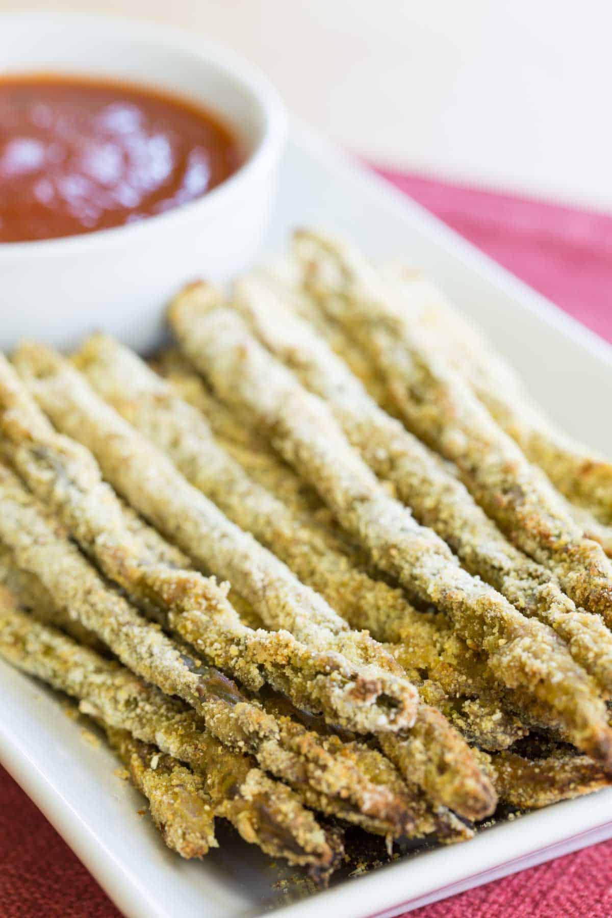 Asparagus spears with a cornmeal breading on a rectangular plate with a bowl of tomato sauce for dipping.