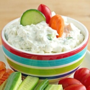 Creamy cucumber dill dip in a striped bowl with a slice of carrot and cucumber and a grape tomato stuck in the top of it.