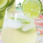 A stemless wine glass filled with a white wine cocktail garnished with cucumber and lime slices on a floral placemat with text overlay that says "Cucumber Gimlet White Sangria".