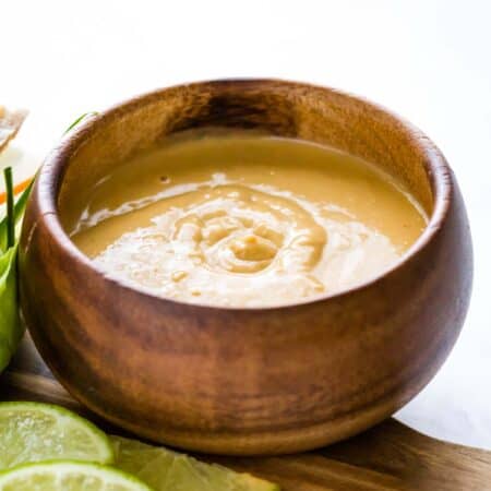 A wooden bowl of peanut sauce on a wooden board with lime slices next to it.