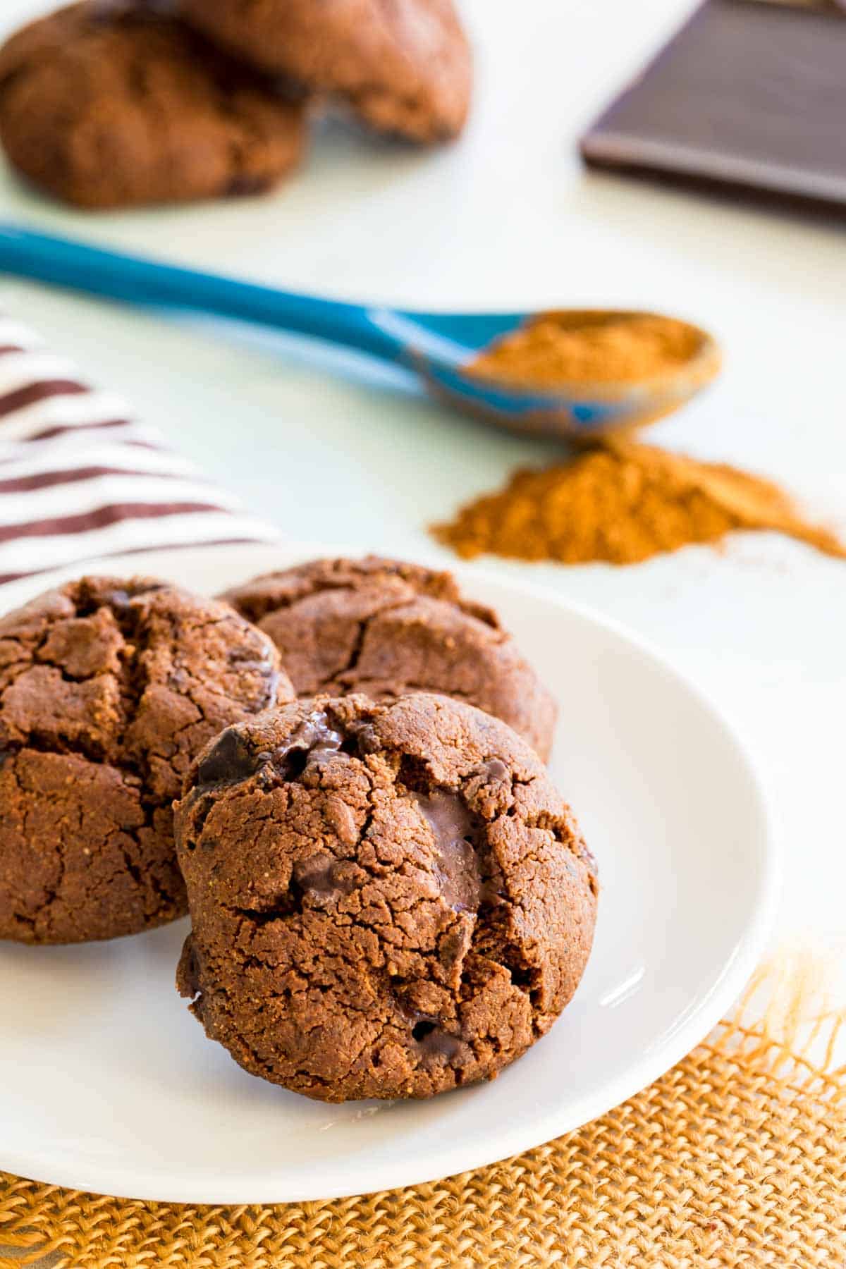 Three Mexican hot chocolate cookies on a plate with chocolate bars and a spoon of spices in the background.