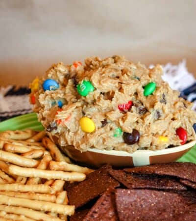 A football-shaped dough of edible monster cookies dough on a plate with pretzel sticks and chocolate tortilla chips.