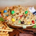 A football-shaped bowl of edible cookie dough with M&M's with text overlay that says "Flourless Monster Cookie Dough Dip".
