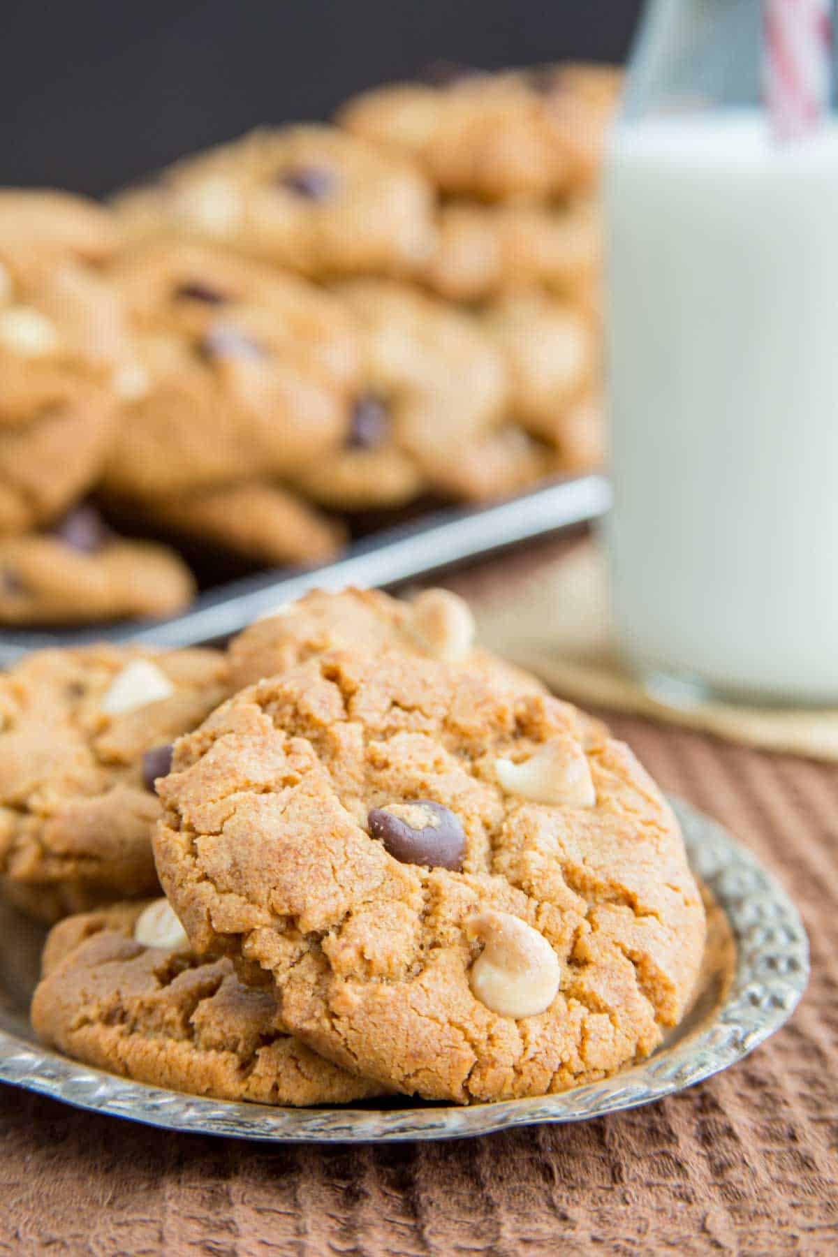 Three peanut butter chocolate chip cookies on a small oval silver plater with a glass bottle of milk and more cookies piled in the background.