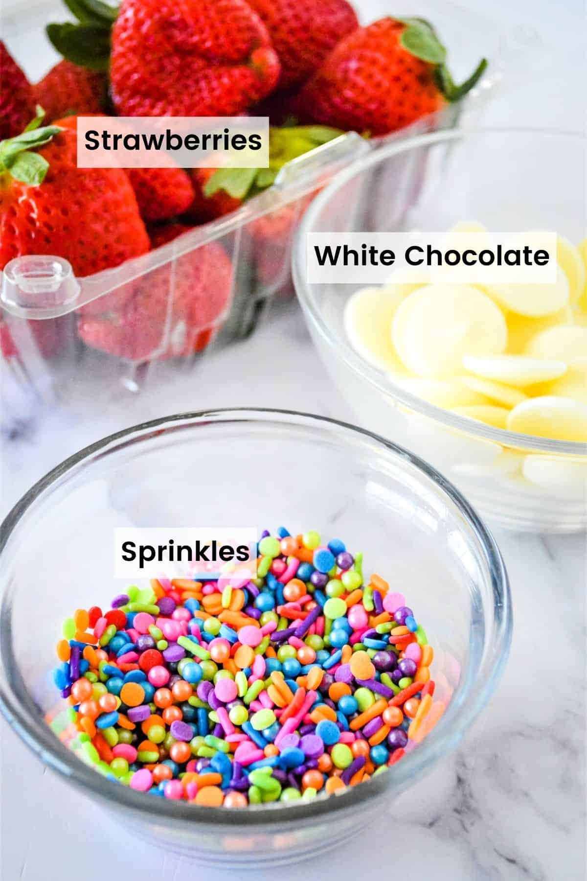 A container of fresh strawberries, a bowl of rainbow sprinkles, and a bowl of white chocolate candy melts