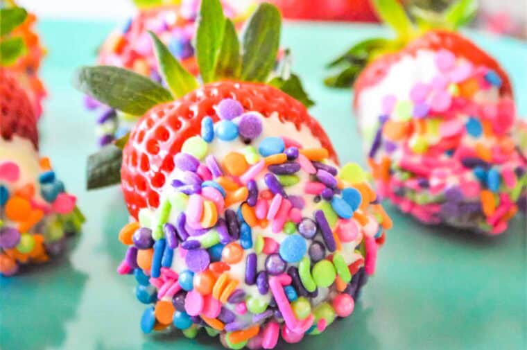 Rainbow sprinkles covering a white chocolate dipped strawberry on a turquoise plate.