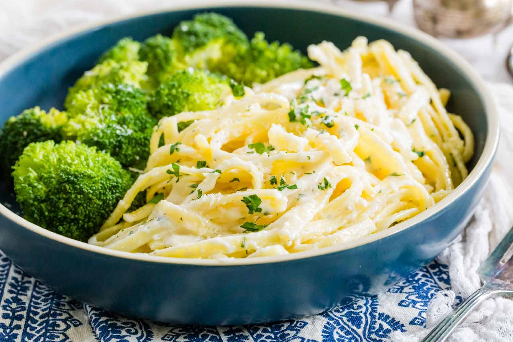 Gluten-Free Fettuccine Alfredo is shown in a bowl topped with parsley with broccoli alongside.