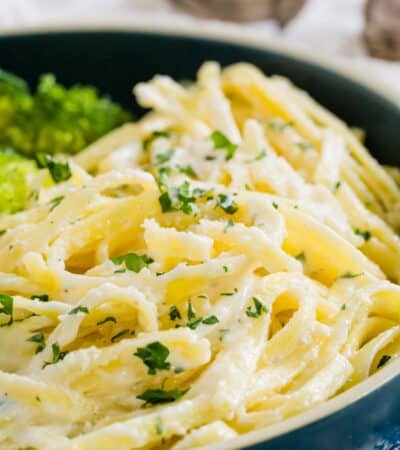 Gluten-Free Fettuccine Alfredo is shown in a bowl topped with parsley with broccoli alongside.