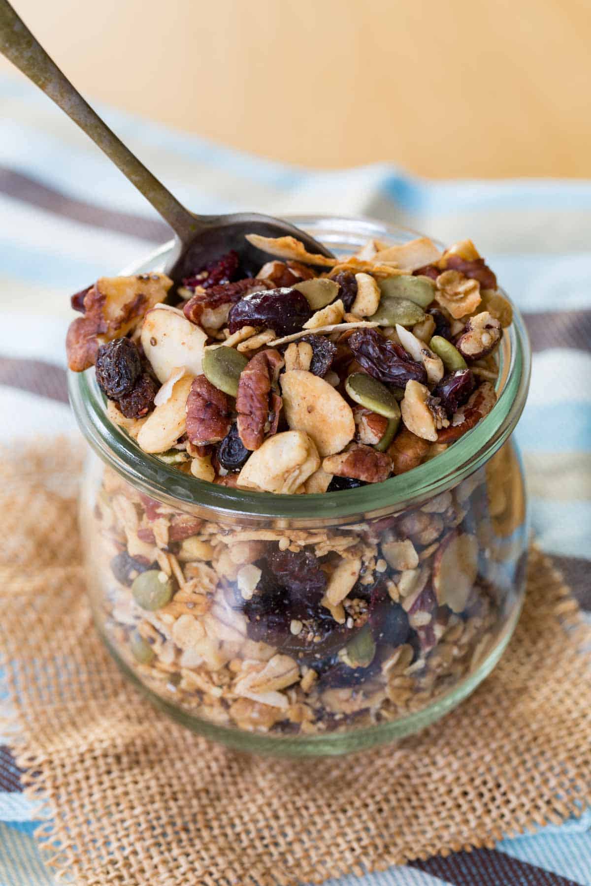 A spoon in a jar of gluten free granola with lots of nuts, seeds, and dried fruit.