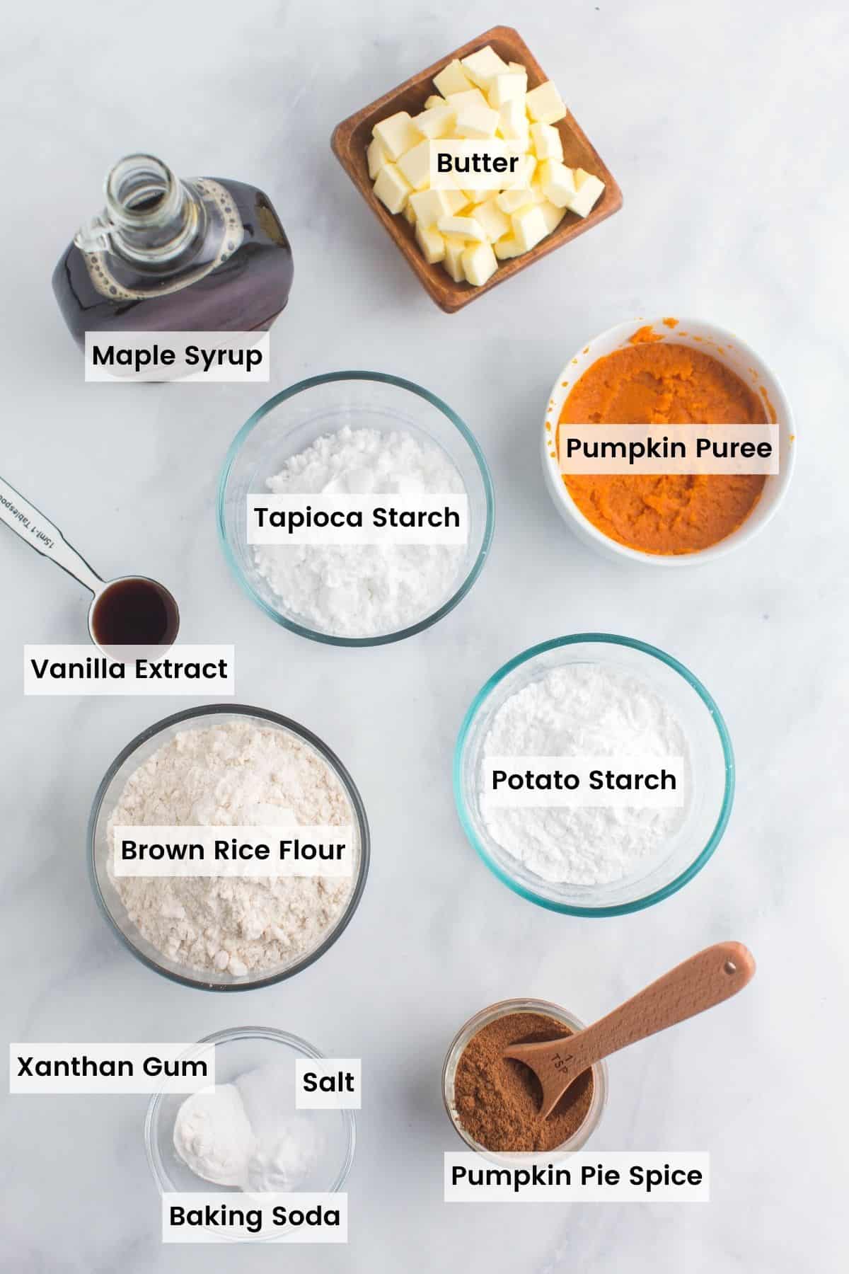 bowls of brown rice flour, tapioca starch, potato starch, salt, baking powder, baking soda, pumpkin puree, pumpkin pie spice, and cubed butter plus a bottle of maple syrup and a measuring spoon of vanilla with text labels.