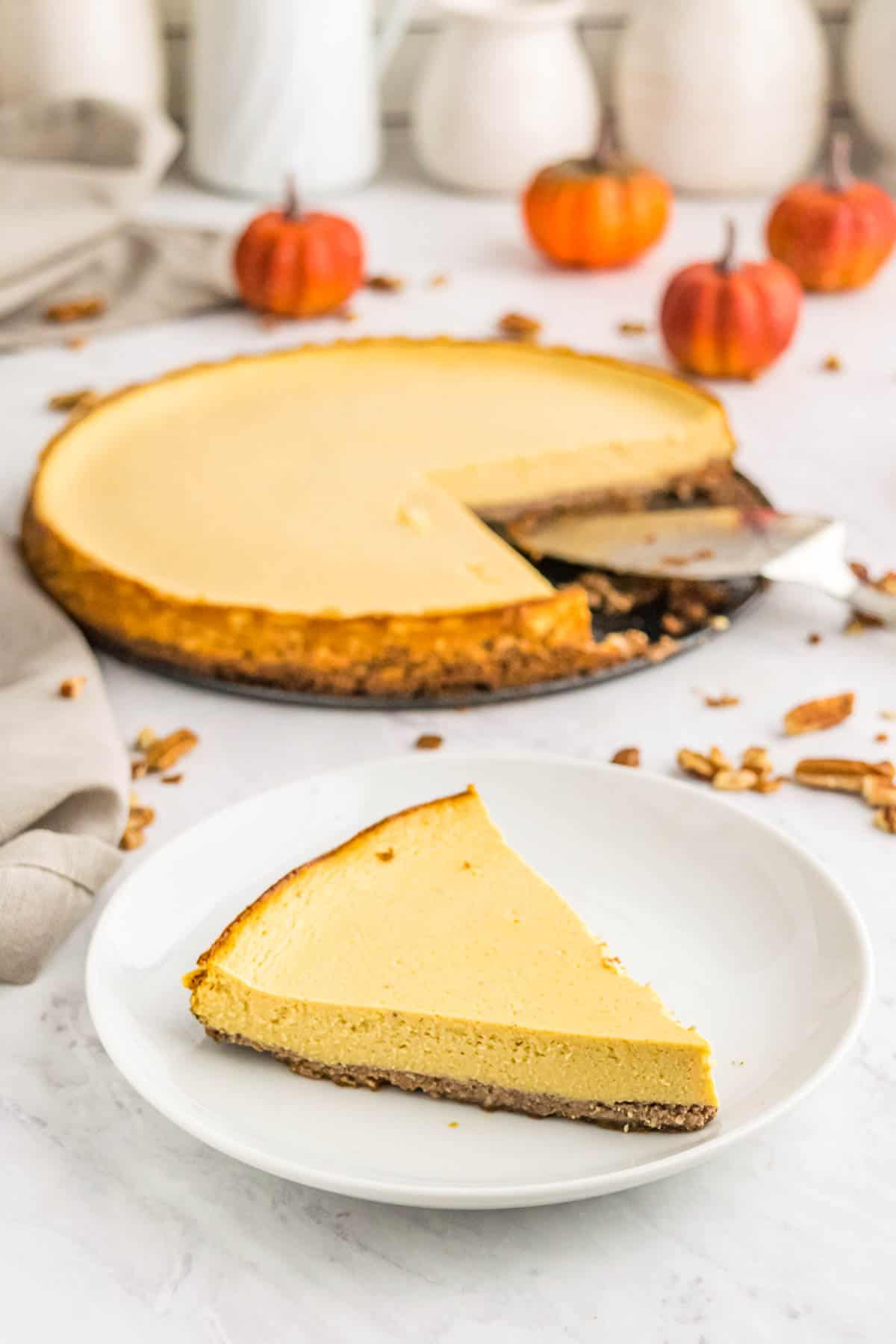 A slice of pumpkin tart on a white plate in front of the rest of the tart with chopped pecans and mini pumpkin decorations scattered around.