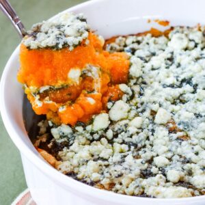 A spoon lifting a scoop of butternut squash casserole with sage pesto and gorgonzola out of the white baking dish.