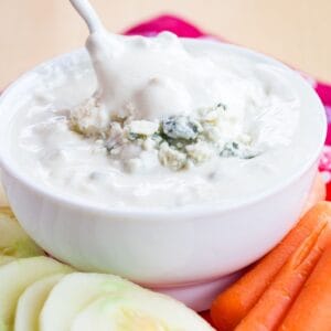 A spoon dipping into a bowl of blue cheese dip topped with crumbles of blue cheese.