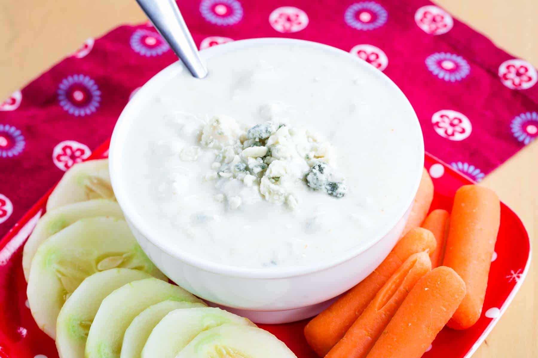 A spoon dipped into a bowl of Greek yogurt blue cheese dip surrounded by carrots and cucumbers.