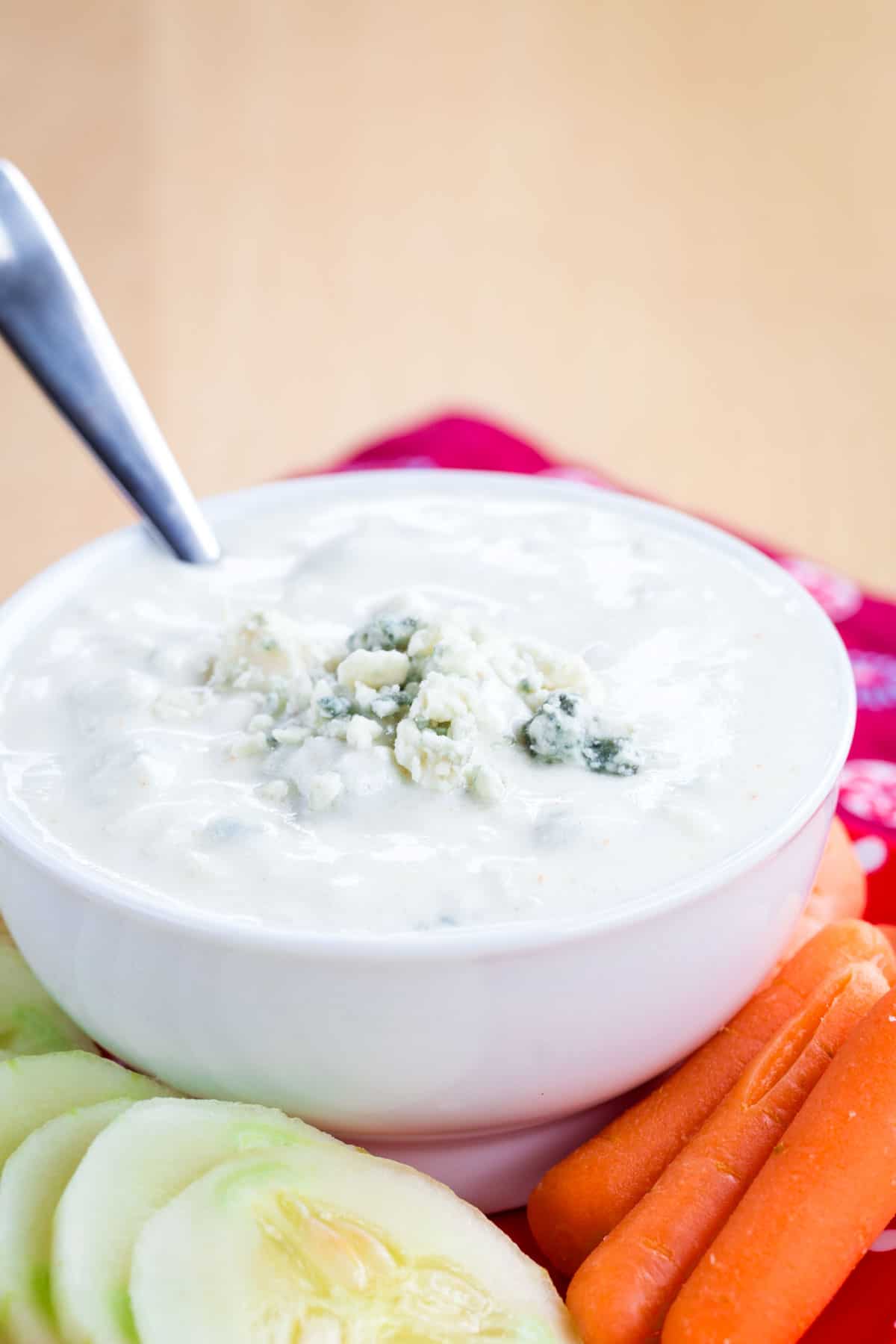 A spoon resting in a bowl of Greek yogurt blue cheese dip surrounded by carrots and cucumber slices.