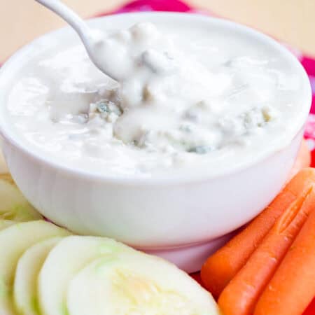 A spoon dipped into a bowl of Greek yogurt blue cheese dip surrounded by veggies.