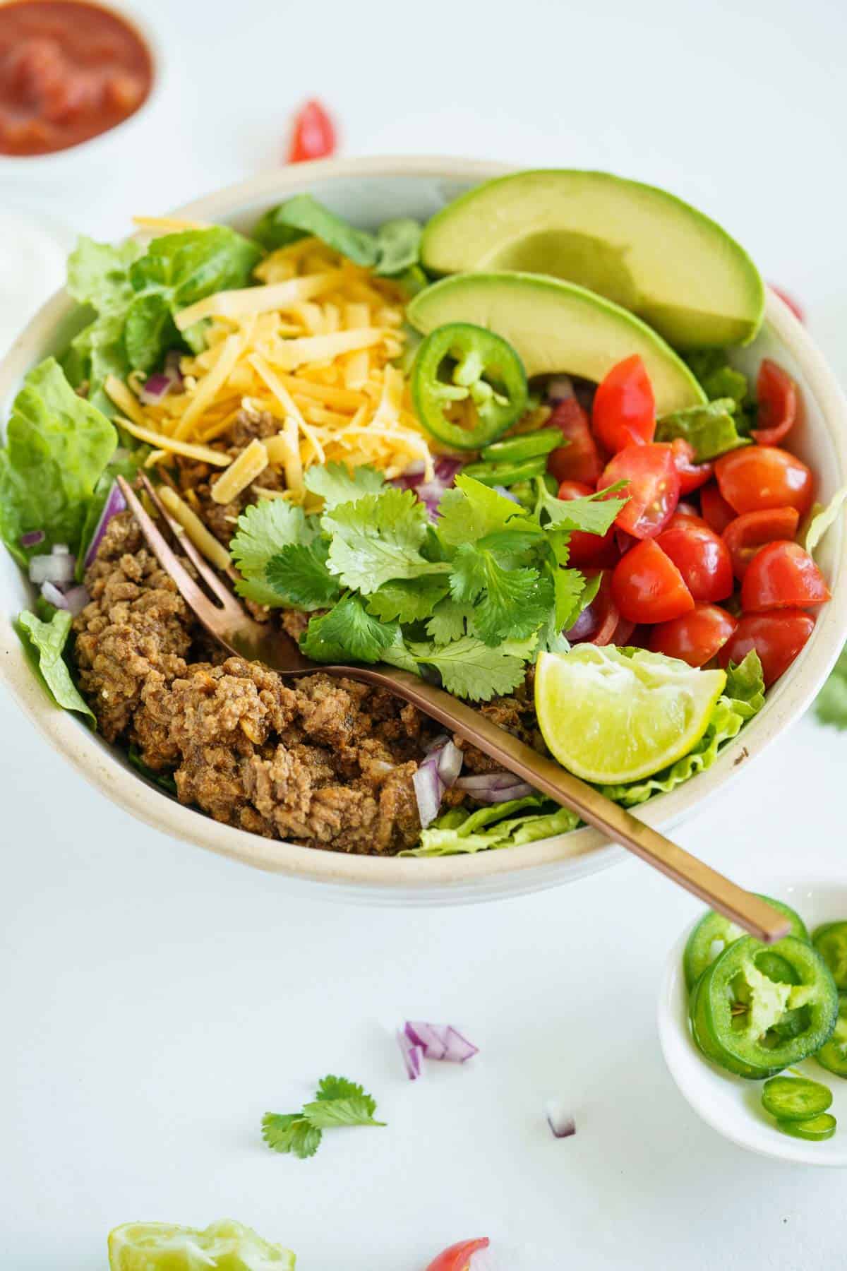 A ground beef taco salad in a bowl with some of the toppings scattered on the table and a small bowl of jalapeno slices next to it.