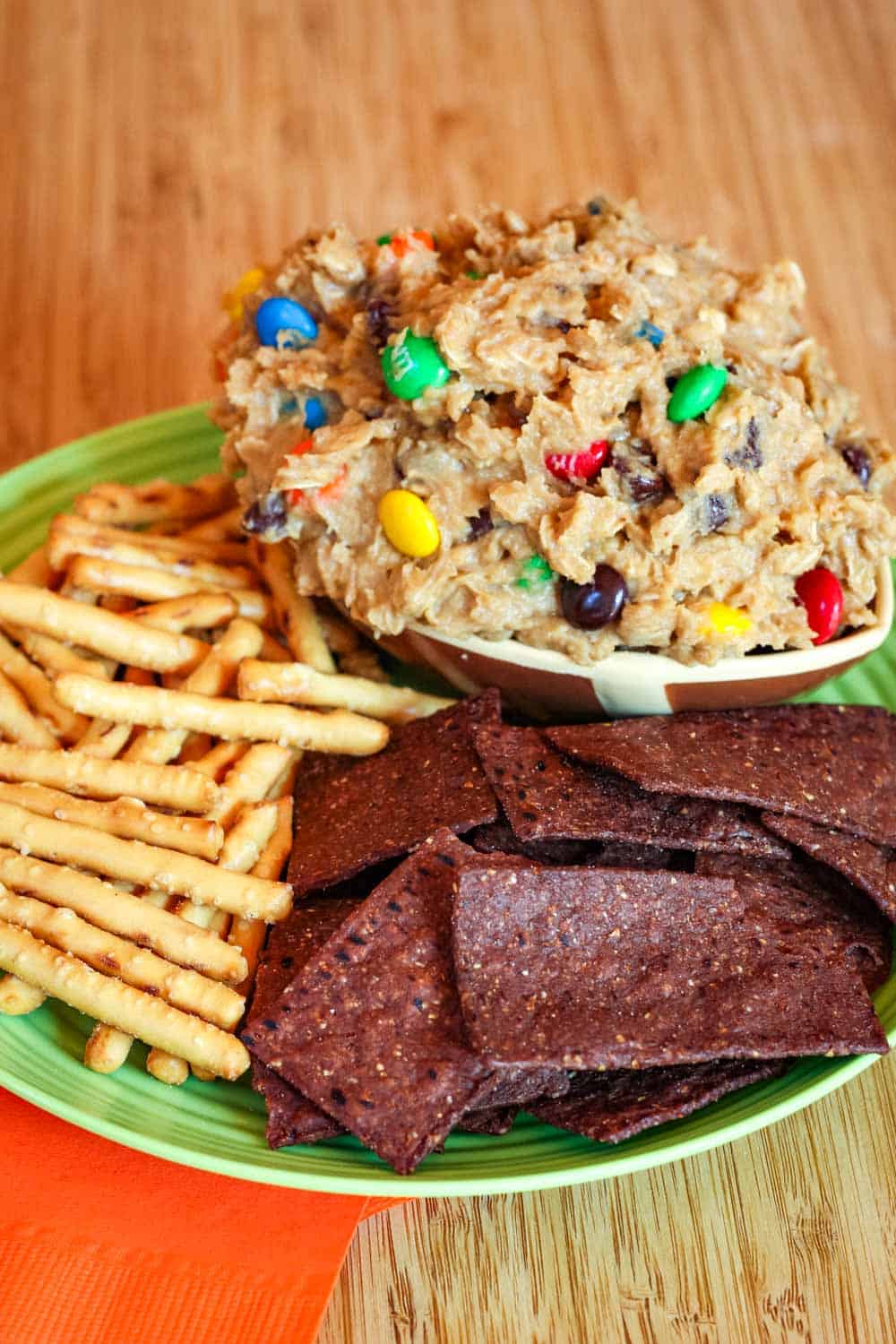 Chips and pretzels on a plate with a bowl of edible cookie dough with oats and M&M's.