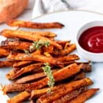 A plate of sweet potato fries garnished with fresh herbs next to a bowl of ketchup with text overlay that says "Baked Sweet Potato Fries".