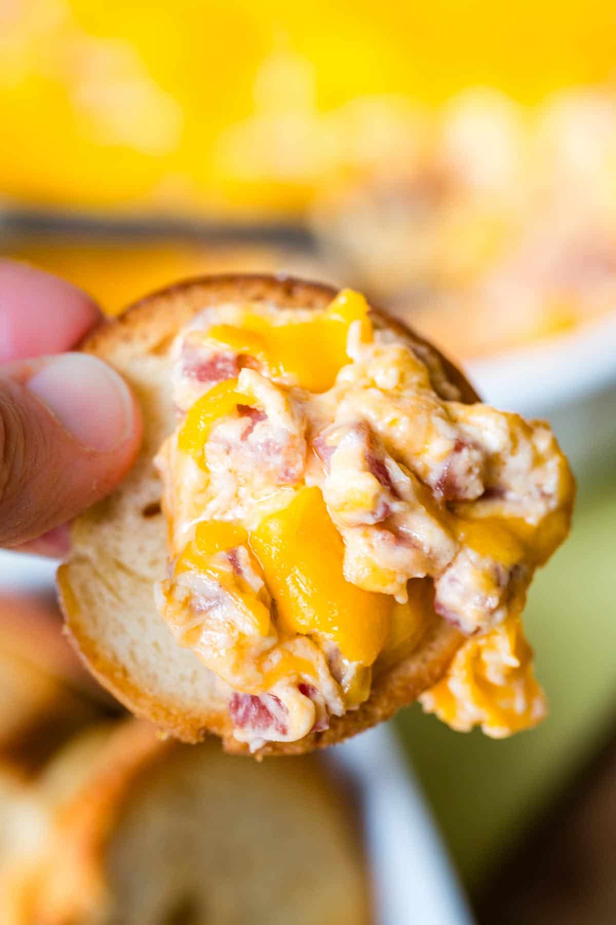 A finger holding a bagel chip with some of the cheesy dip on it.