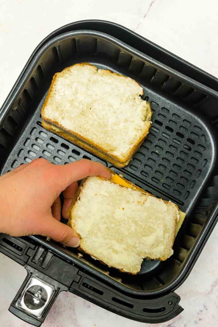 A hand places a piece of buttered bread on top of a grilled cheese sandwich in an air fryer.