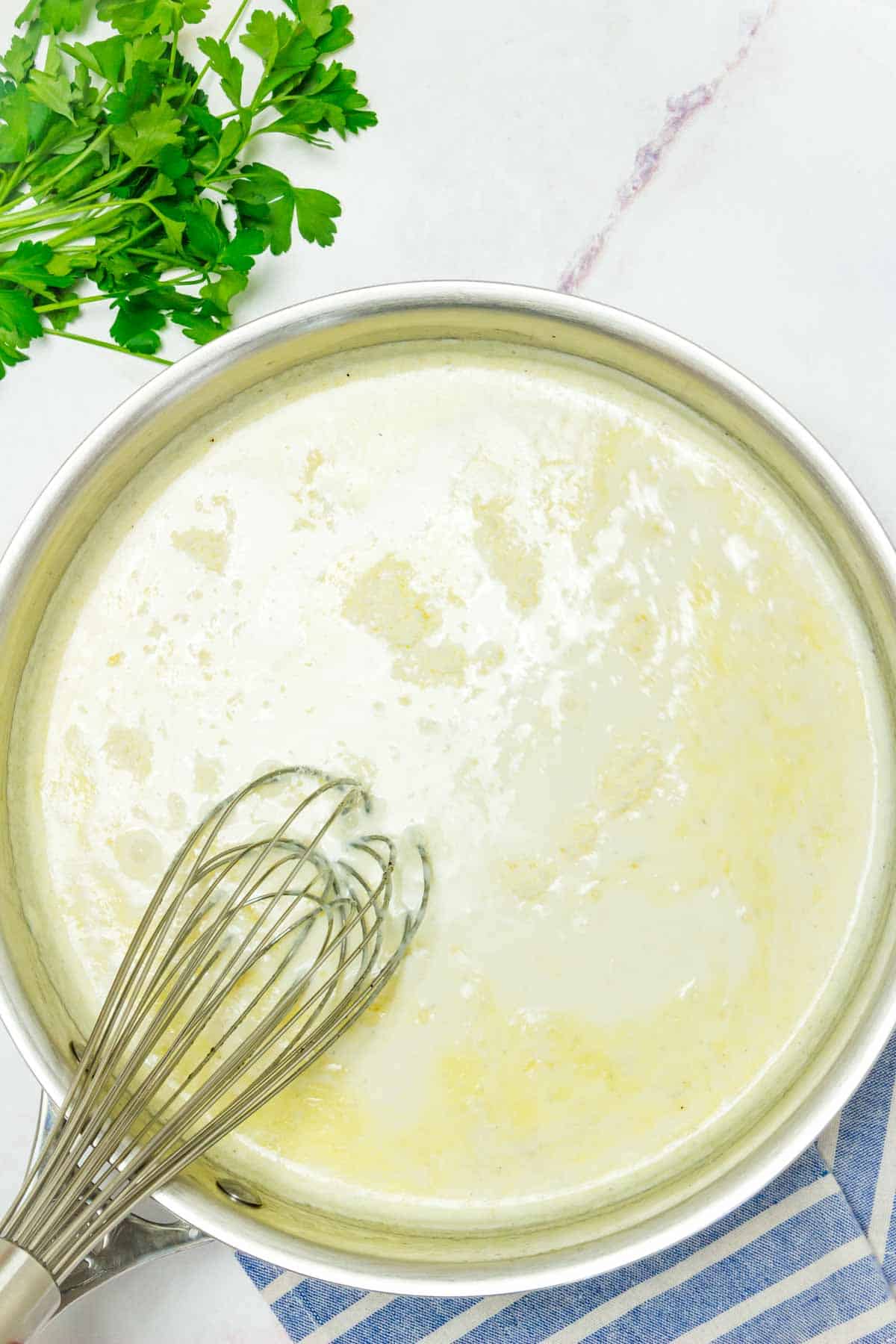 A whisk helps combine a pot of alfredo sauce with a bunch of parsley on the side.