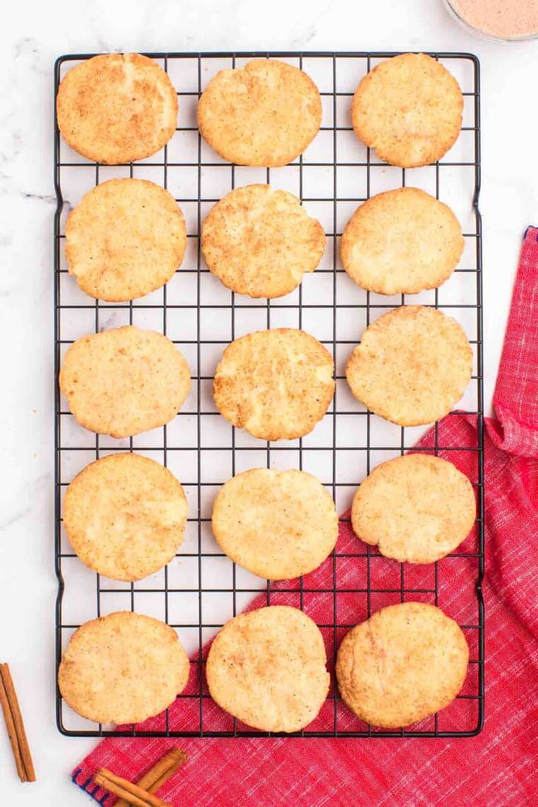 24 snickerdoodles scattered on a cooling rack with a bowl of cinnamon sugar on the side.