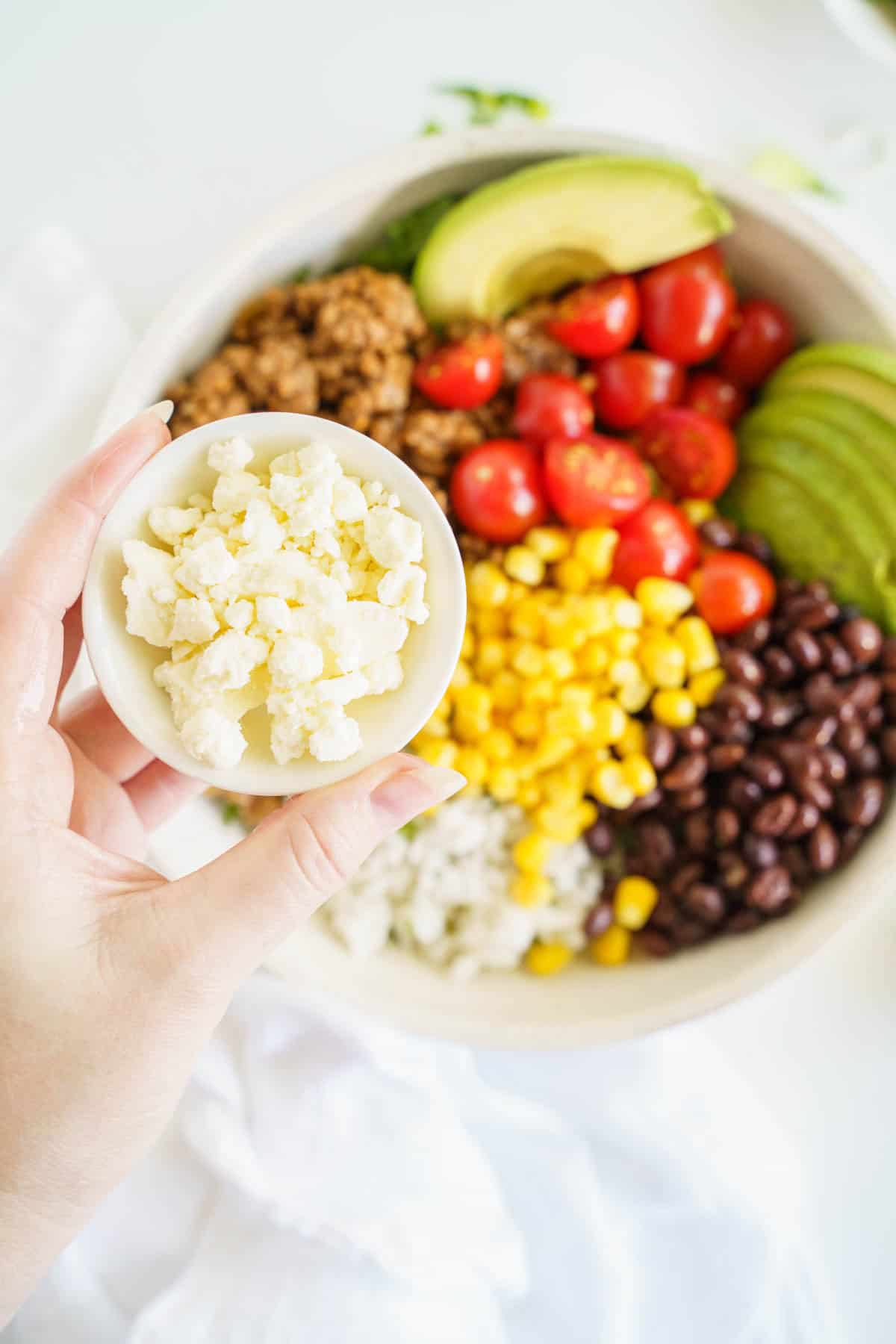 A Small Dish of crumbled queso fresco Being Held Over a Burrito Bowl.