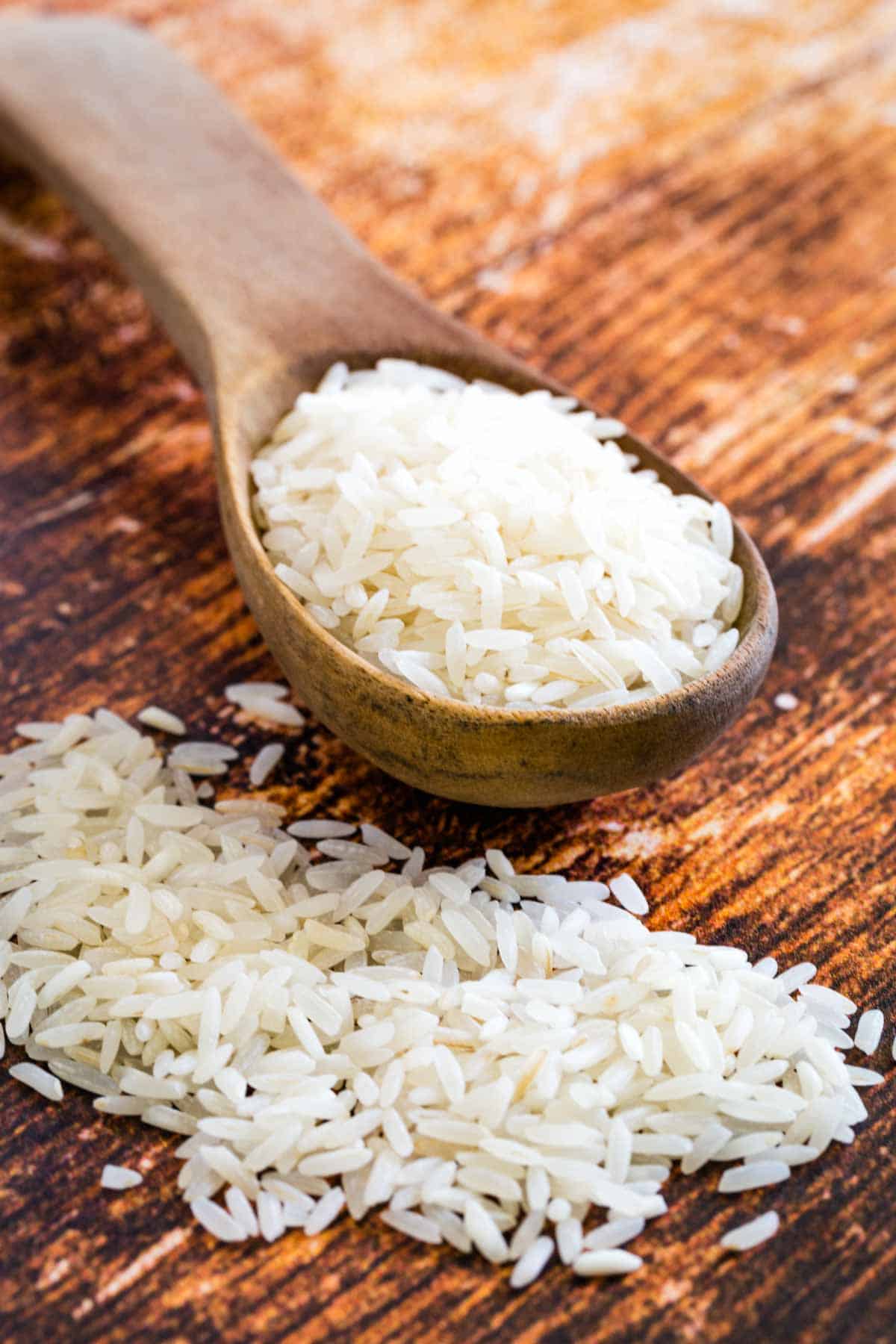 A large wooden spoonful of jasmine rice on a wooden countertop, with scattered rice grains.