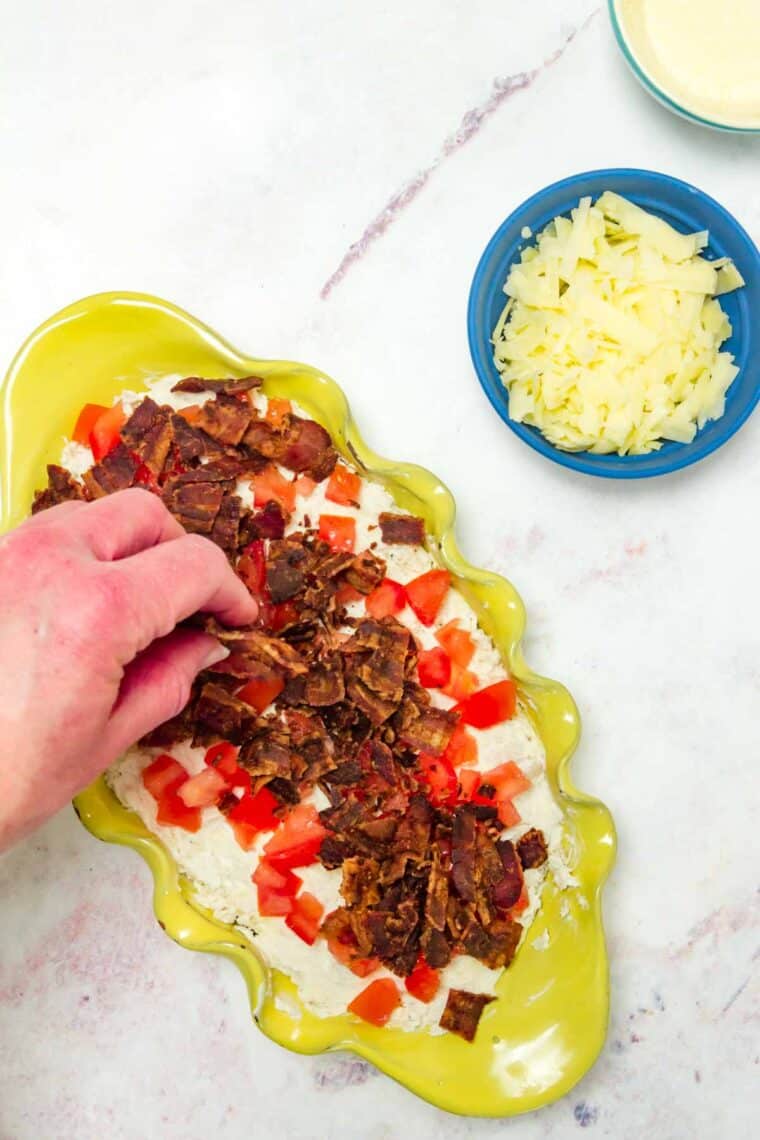 A hand sprinkling cooked bacon over the layer of tomato on top of the dip mixture in the baking dish.