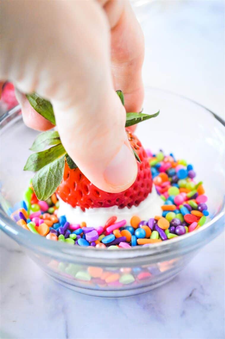 Dipping a white chocolate covered strawberry into a bowl of rainbow sprinkles.
