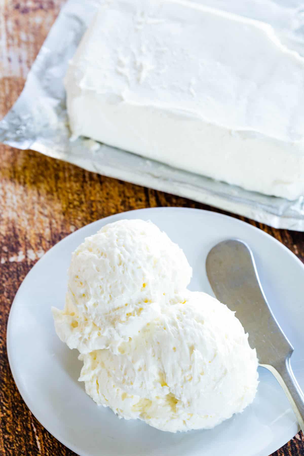 Scoops of whipped cream cheese on a whit plate with a small spread knife and a block of cream cheese blurred in the background.
