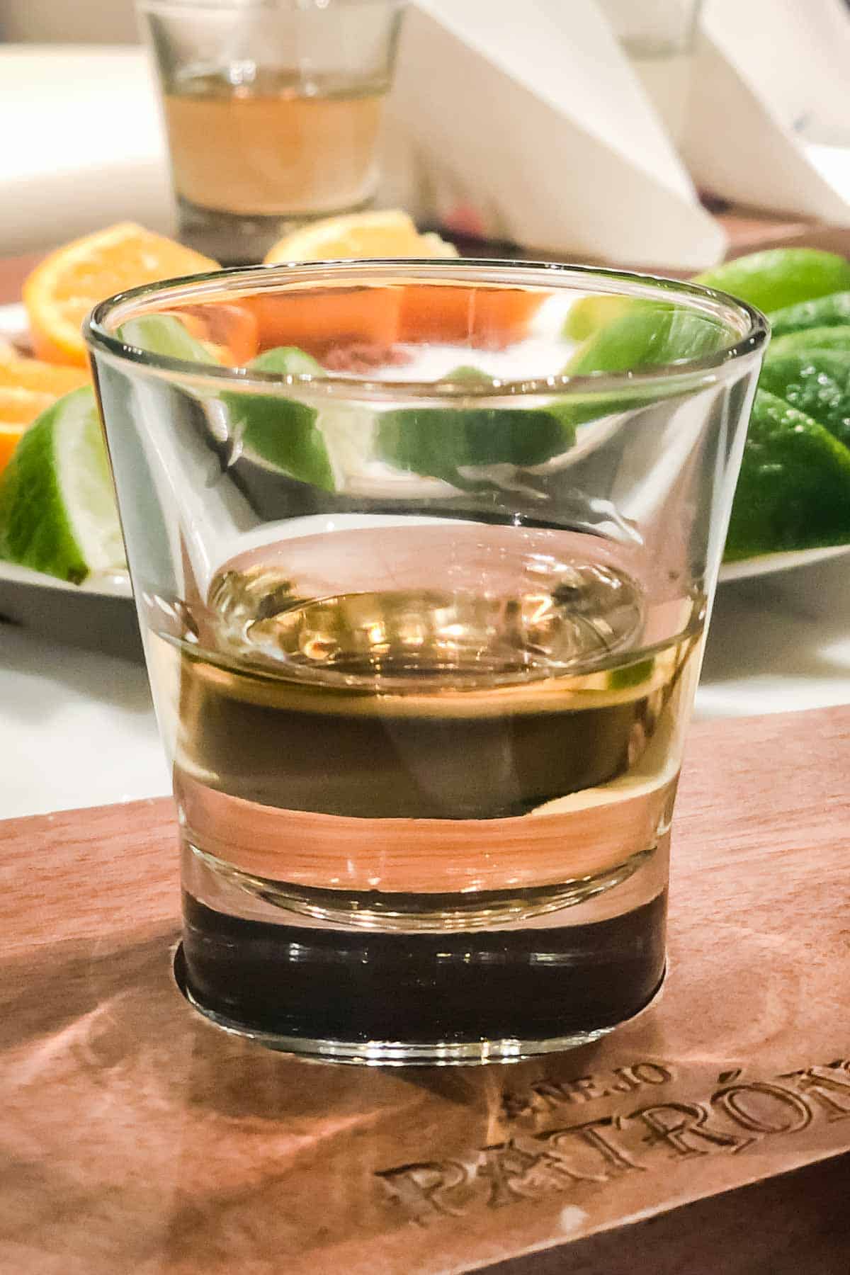 A shot glass of tequila on a wooden board with a plate of lime and orange wedges behind it.
