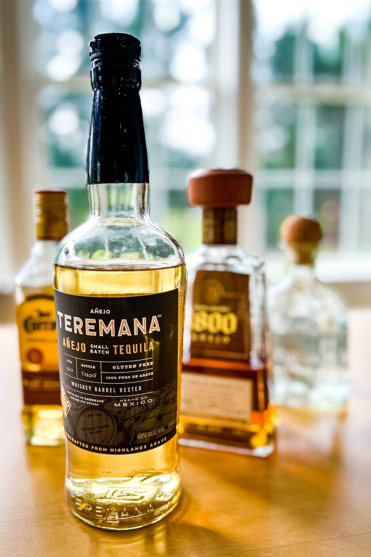A bottle of Teremana tequila on a wood table with a few more bottles of different brands blrured in the background.