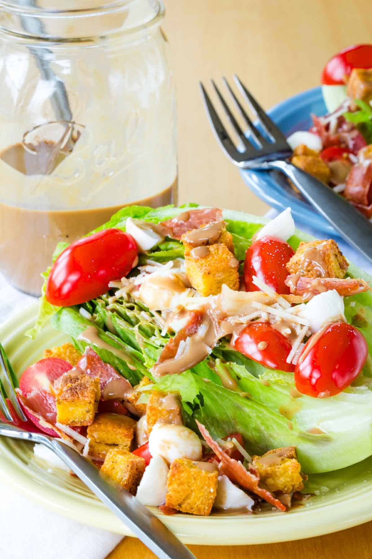 A half of a romaine heart topped with tomatoes, mozzarella, prosciutto, and croutons with a jar of dressing behind it.