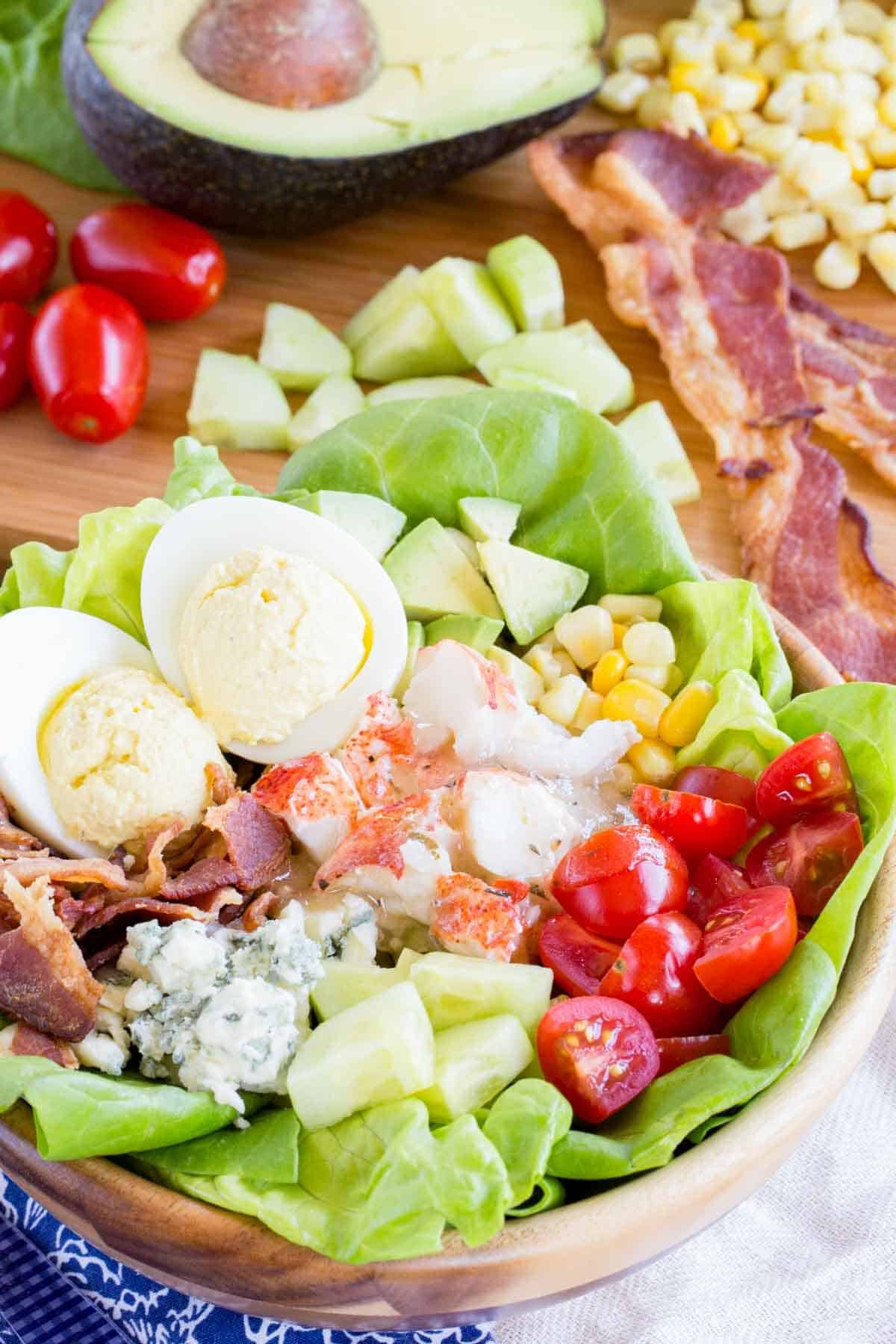 A salad in a wooden bowl with lobster tail meat, eggs, bacon, tomatoes, corn and avocado.