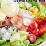 A bowl of salad topped with lobster meat, cucumbers, tomatoes, cheese, corn, bacon, avocado, and eggs with text overlay that says "Lobster Cobb Salad".