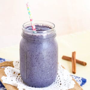 A blueberry smoothie in a glass jar with a straw and cinnamon sticks on the table in the background.