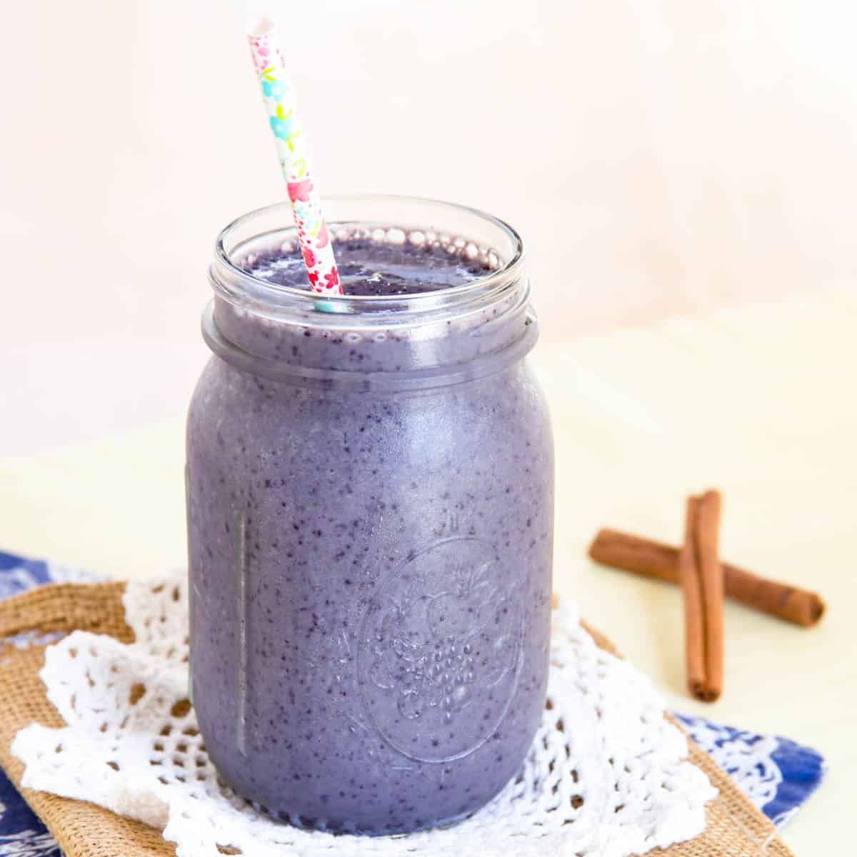 A blueberry smoothie in a glass jar with a straw and cinnamon sticks on the table in the background.