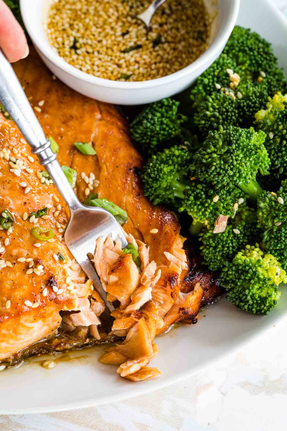 A fork flaking apart broiled maple glazed salmon next to a bowl of maple glaze and broccoli on a plate.