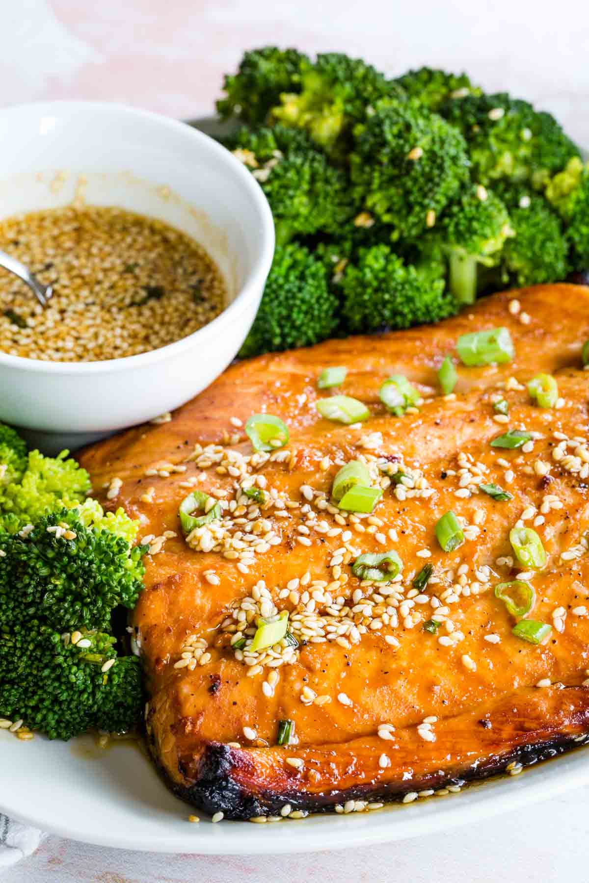 Closeup of a filet of maple glazed salmon topped with sesame seeds and scallions on a plate next to a bowl of marinade and steamed broccoli.