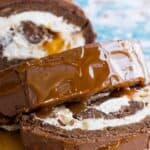 A sliced chocolate roll cake filled with whipped cream and toped with ganache and caramel on a wooden platter with text overlay that says "Milky Way Caramel Swiss Roll".