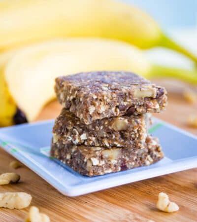 Three Banana Bars stacked on a square blue plate with chopped walnuts around it and bananas in the background.