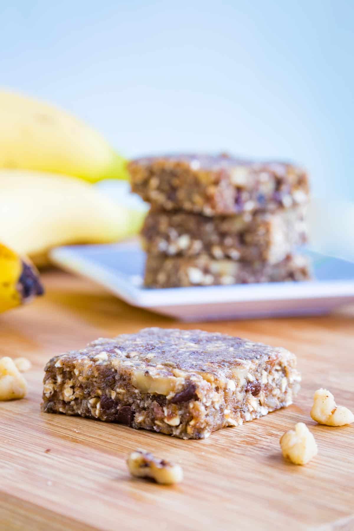 One banana snack bar with chopped walnuts around it and a stack of three energy bars on a plate in the background.