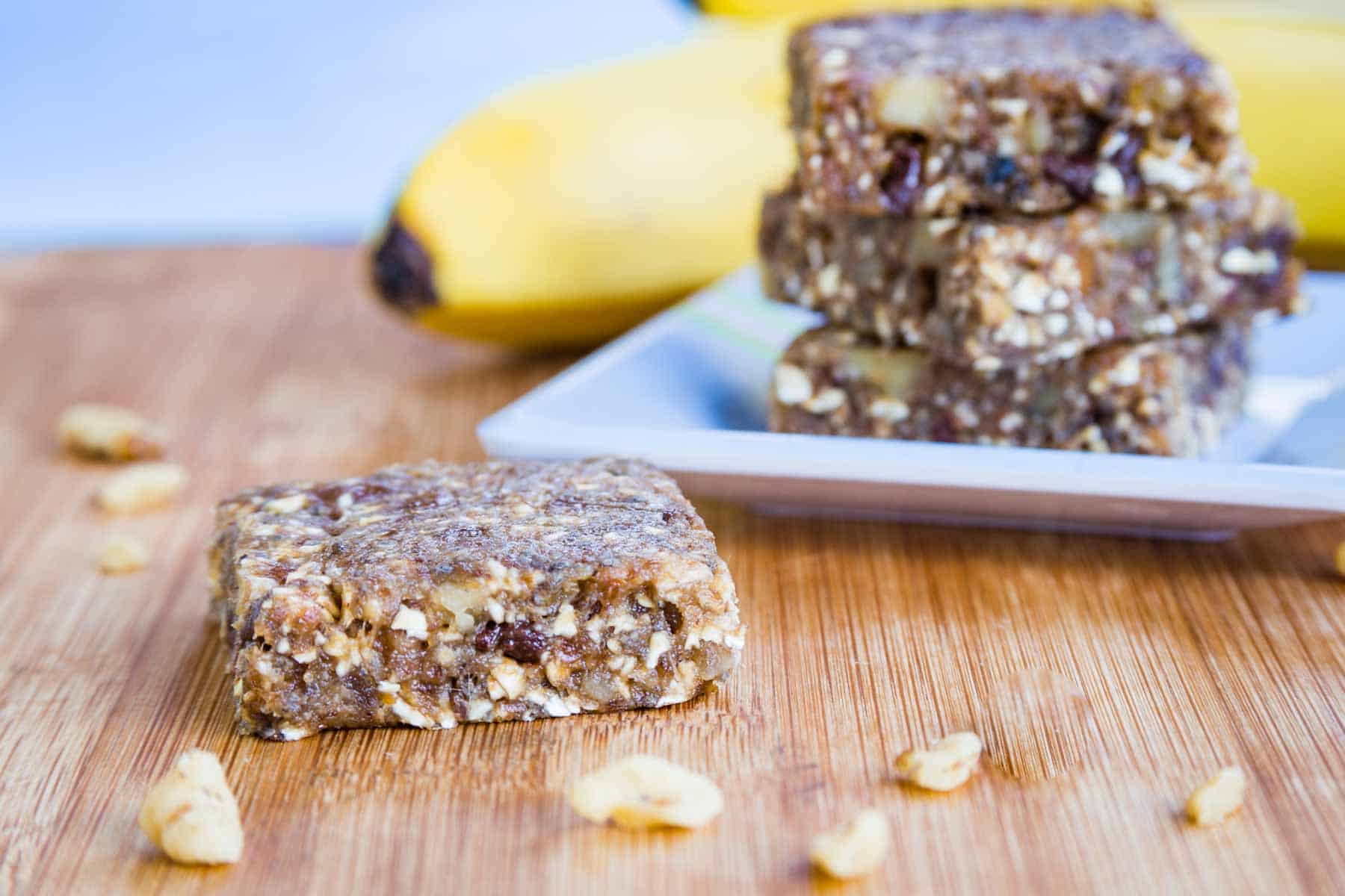 A Banana Nut Bar on a cutting board with walnuts around it and a plate of more bars and bananas in the background.