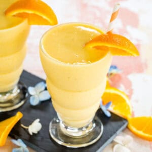 An orange smoothie in a glass with a striped straw and an orange wedge.