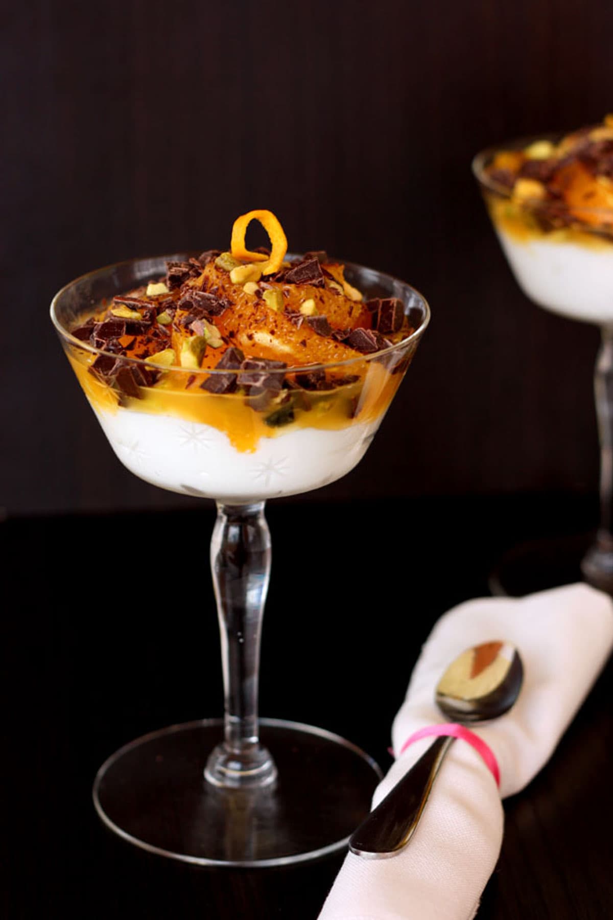 A stemmed dessert glass filled with yogurt, orange slices, dark chocolate chunks, and pistachios with a rolled cloth napkin and spoon next to it.