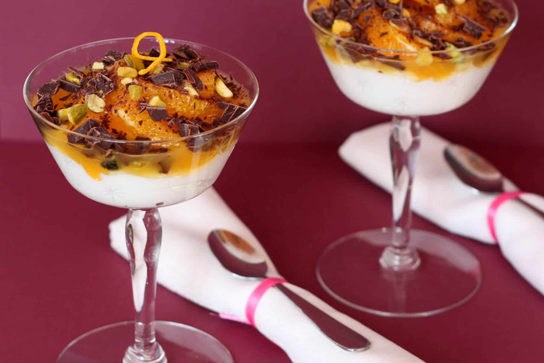Two stemmed dessert glasses filled with yogurt, orange slices, dark chocolate chunks, and pistachios with rolled cloth napkins and spoons next to them.