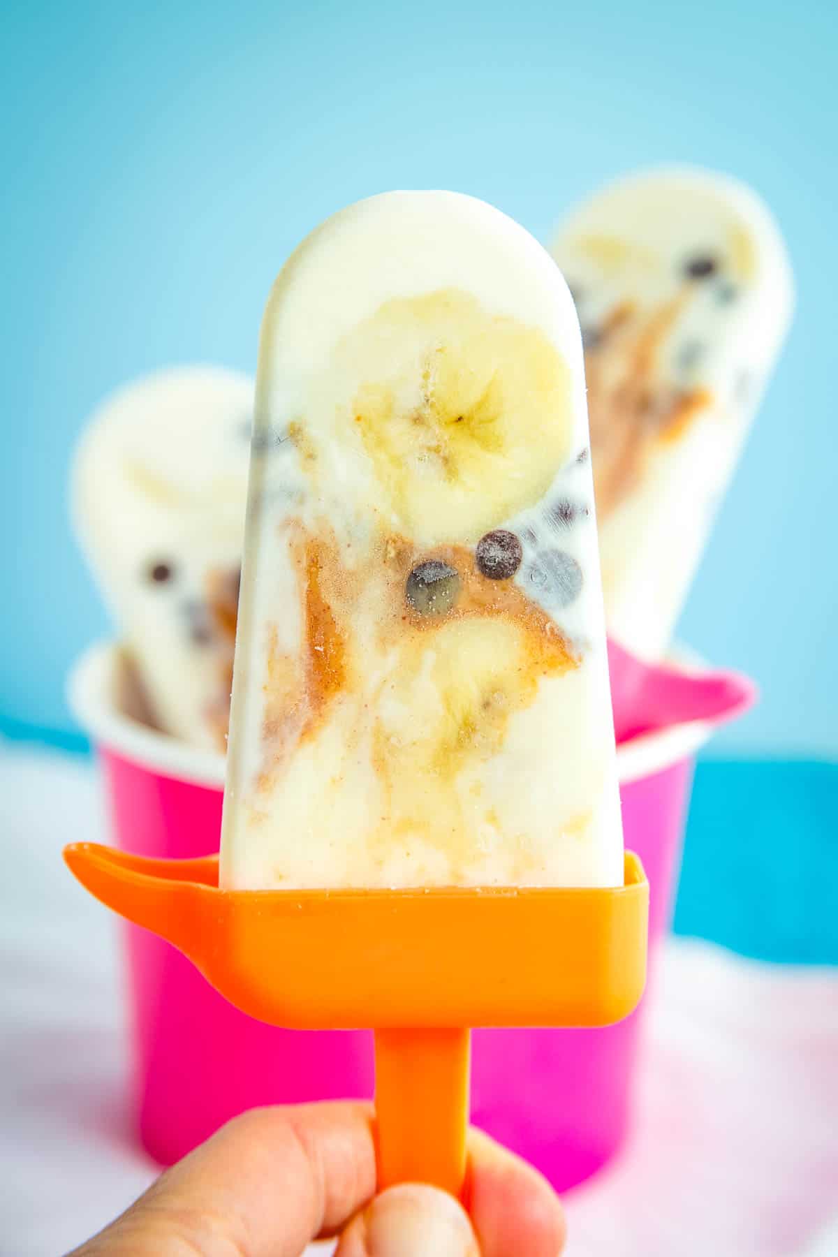 A hand holding a frozen yogurt popsicle with banana slices, chocolate chips, and swirls of peanut butter on an orange handle.