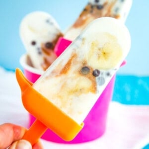 A frozen yogurt pop with banana slices, mini chocolate chips, and a swirl of peanut butter on an orange handle.